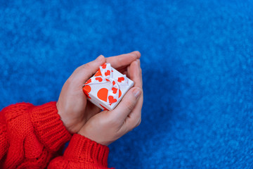 Hands of a girl in a red sweater holding a box with a gift with a heart on a blue glitter background.