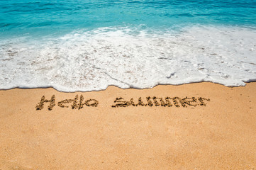 Hand written text hello summer on the golden beach sand with coming waves
