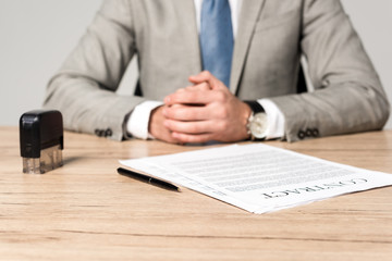 cropped view of businessman sitting at desk near contract, stamp and pen isolated on grey