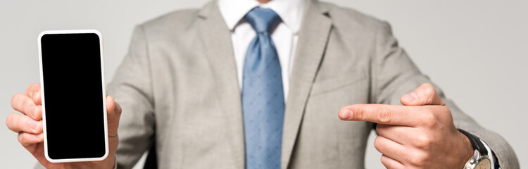 cropped view of businessman pointing with finger and smartphone with blank screen isolated on grey, panoramic shot