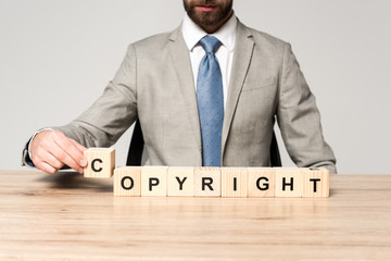 cropped view of businessman sitting at desk near wooden cubes with word copyright isolated on grey