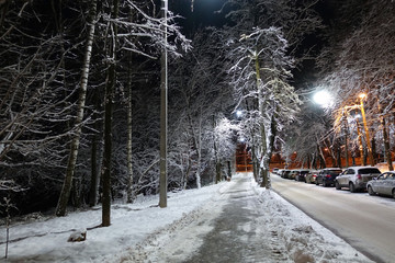 Night street in winter consecrated by lanterns. Paths, trees, cars covered in snow.