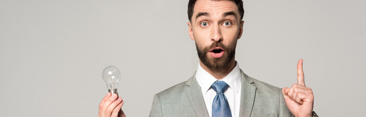 panoramic shot of excited businessman holding light bulb and showing idea sign isolated on grey