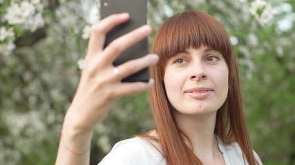 A girl makes selfie in the garden. An attractive red-haired woman smiles making selfi using a mobile phone in a cherry orchard. The concept of using gadgets for a healthy lifestyle.