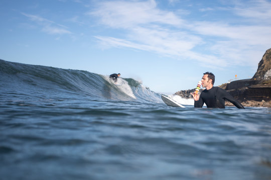 Guy Eating An Ice Cream Cone With Different Flavors While A Surfer Catches A Wave In The Background