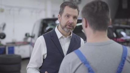 Close-up portrait of displeased Caucasian man talking with unrecognizable auto mechanic. Adult confident businessman unsatisfied with repair shop service. Warranty, car business.