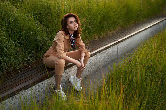 Young beautiful red haired gen z girl wearing slim fitted women beige khaki coverall overall denim jumpsuit or flight suit and white chunky platform sneakers sitting on a long park bench in high green