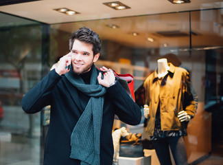 Attractive young man with a beard who has bags in one hand is talking on the phone while shopping in the city.