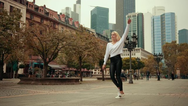 Young girl dancer ballerina blonde dancing on street of modern city metropolis, dressed in white sweater and black tight pants