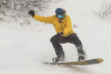 A snowboarder in snow going downhill amd jumping. Boarder with yellow clothing with black trousers and orange board riding and jumping over snow on a cloudy day.