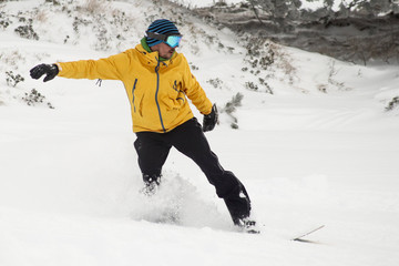 A snowboarder in snow going downhill amd jumping. Boarder with yellow clothing with black trousers and orange board riding and jumping over snow on a cloudy day.