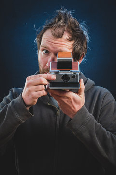 A young man with beard is preparing to take a picture with vintage photo camera. Frontal shot of an instant photographer in a studio with black background and blue lighting.