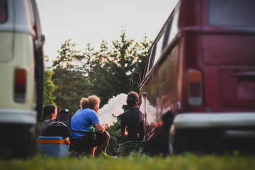 A group of people sitting enjoying a picninc and a smoke with old time vans trucks parked behind...