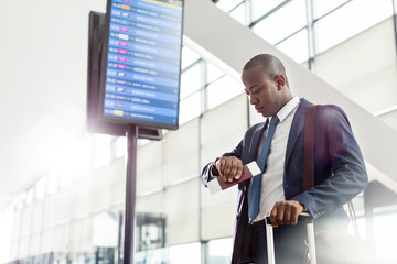 Businessman checking the time on wristwatch below arrival departure board at airport