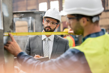 Manager watching steel worker with tape measure in factory
