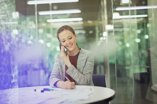 Smiling businesswoman talking on cell phone taking notes in conference room