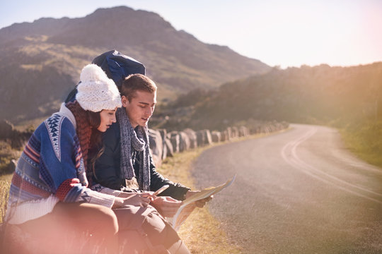 Young couple backpacks hiking, resting at sunny, remote roadside map smart phone