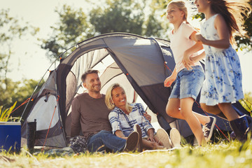 Parents watching happy daughters running around sunny campsite tent
