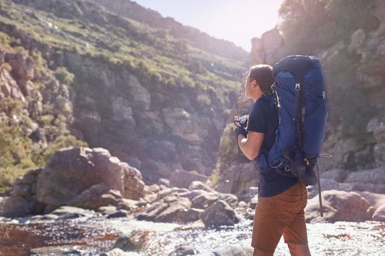 Young man backpack hiking photographing camera below sunny, remote cliffs