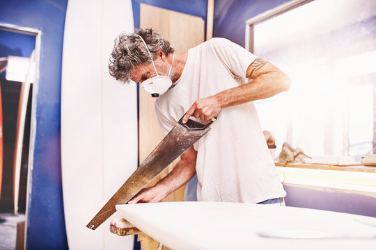 Male surfboard designer wearing protective mask using saw in workshop - Powered by Adobe
