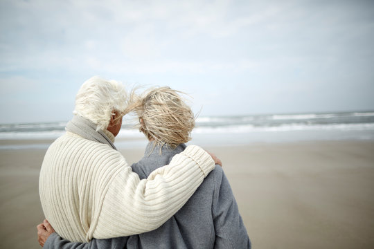 Pensive senior couple hugging looking at ocean view on windy winter beach