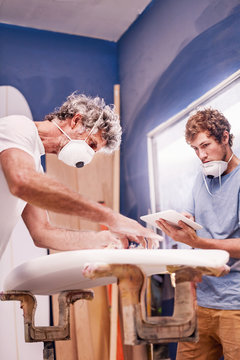 Male surfboard designers wearing protective masks sanding surfboard in workshop