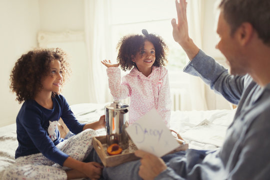 Father receiving Father&sbquo;&Auml;&ocirc;s Day card from daughters high-fiving on bed