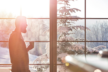 Man drinking coffee at sunny window with view of snowy trees