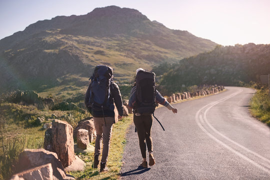 Young couple with backpacks hiking along sunny, remote roadside