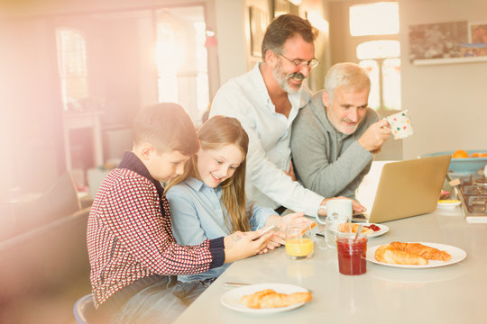 Male Gay Parents Children Using Laptop Cell Phone, Eating Breakfast In Kitchen