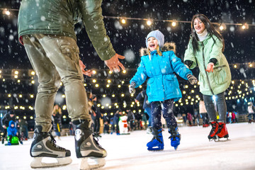 Skating rink. Happy family on the ice rink. Mom and dad teach daughter to skate. - obrazy, fototapety, plakaty