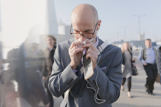 Businessman blowing nose tissue, holding cell phone headphones on urban pedestrian bridge