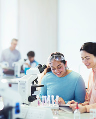 Smiling female science teacher and girl student reading textbook at microscope in laboratory classroom