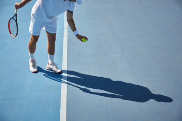 Young male tennis player playing tennis, bouncing tennis ball on sunny blue tennis court