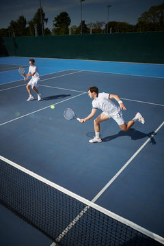 Young male tennis doubles players playing tennis, reaching for the ball on sunny blue tennis court