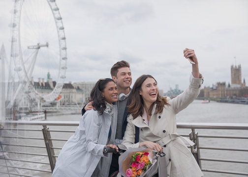 Smiling, happy friends taking selfie selfie stick on bridge near Millennium Wheel, London, UK
