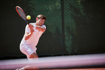 Young male tennis player playing tennis, hitting the ball on sunny tennis court