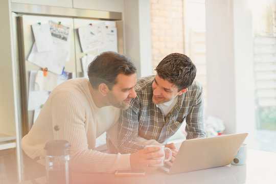 Male Gay Couple Using Laptop And Drinking Coffee In Kitchen