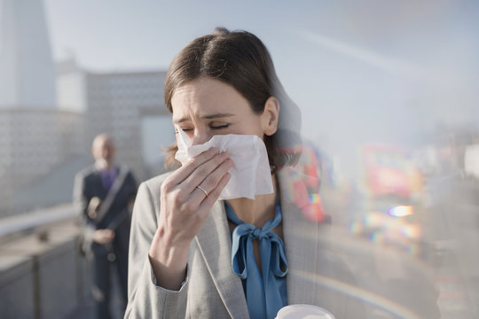 Businesswoman allergies blowing nose into tissue on sunny urban sidewalk