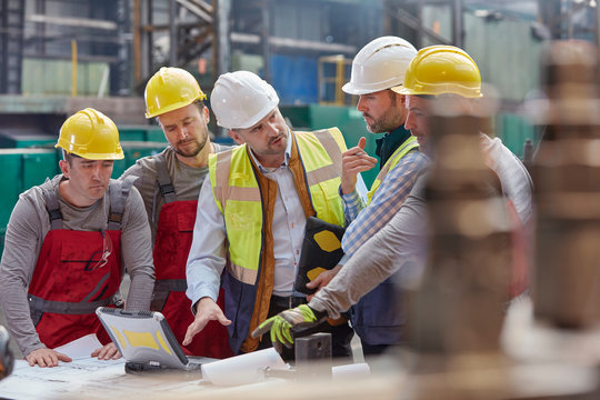 Male foreman, engineers workers at laptop meeting, discussing blueprints in factory