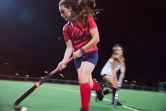 Determined young female field hockey player hitting the ball, playing on field at night
