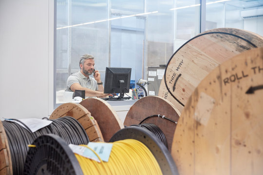 Male supervisor working at computer talking on ell phone behind spools in fiber optics factory
