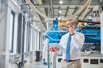 Male supervisor digital tablet talking on cell phone next to plastic industrial molder machinery in factory
