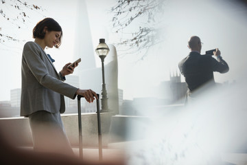 Businesswoman suitcase using cell phone in sunny urban park, London, UK