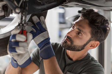 Focused male mechanic fixing wheel underneath car in auto repair shop