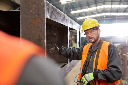 Male worker gesturing, explaining steel part to colleague in factory