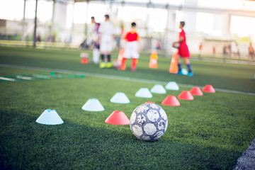 Football and Marker Cone on green artificial turf with blurry kid soccer players are training background. Soccer academy.