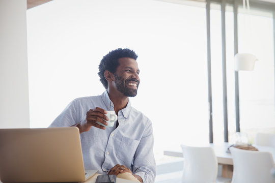 Smiling, enthusiastic man drinking coffee and working at laptop at home