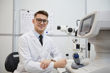 Portrait of male optometrist smiling at camera while posing at workplace by optic equipment, copy...