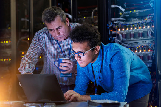 Focused male IT technicians working at laptop in dark server room